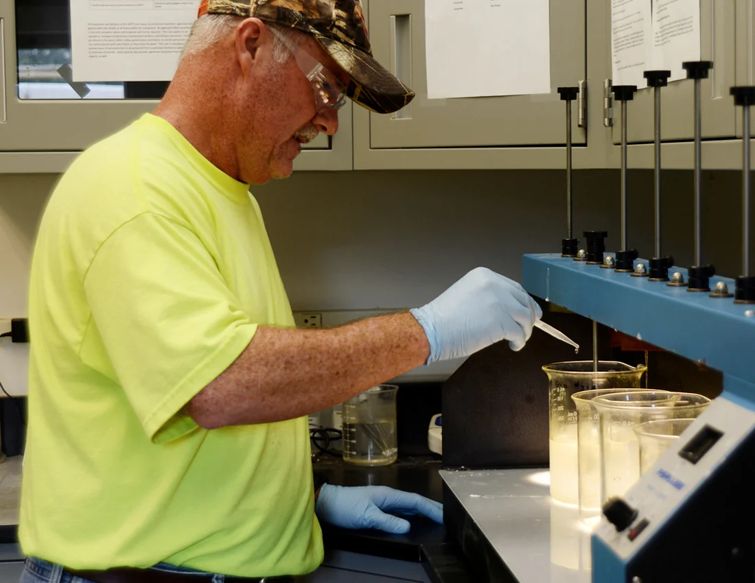 A wastewater operator wearing safety glasses and gloves performing a jar test in a treatment-plant laboratory